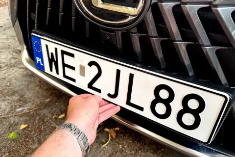 Dłoń poprawia tablicę rejestracyjną auta. Wartość samochodu powyżej 150 tys. zł, widoczna na grillu i zderzaku.