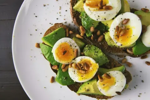 Avokado recept: två rostade brödskivor med avokado, kokt ägg och pinjenötter, strösslade med peppar.