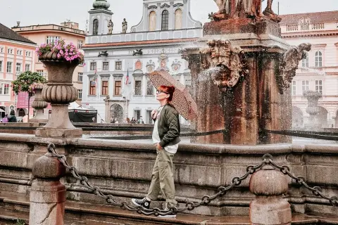 Regenschirm-Schwung am Brunnen in Budweis Stadt. Die Frau genießt die frische Brise, während die historische Kulisse im Hintergrund erstrahlt.