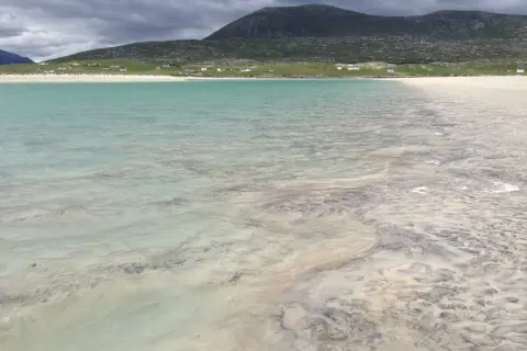 Vackert turkost vatten möter en sandstrand med mönster från tidvattnet. Hus och en grön kulle syns i bakgrunden på Yttre Hebriderna.
