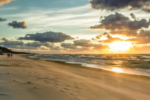 Zachód słońca nad Bałtykiem, plaża i falochron. W oddali widać ludzi spacerujących i kitesurferów. To idealne miejsce na podziwianie atrakcji Jastrzębiej Góry i okolic.