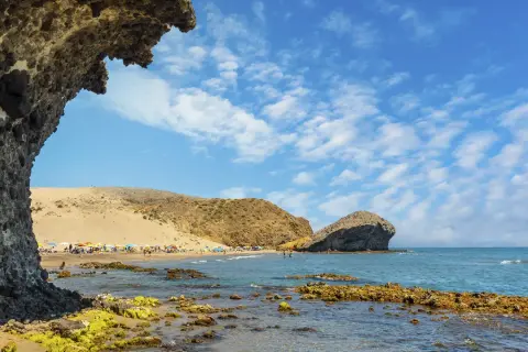 Vista de la playa de San José con rocas cubiertas de algas en primer plano y gente disfrutando del sol bajo sombrillas.