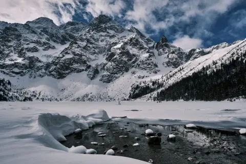 Morskie Oko zimową porą. Majestatyczne, ośnieżone szczyty Tatr górują nad zamarzniętym jeziorem. W oddali widać ludzi spacerujących po lodzie.
