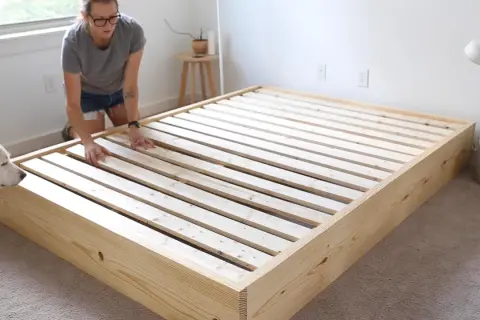 A woman and her dog inspect a newly built DIY platform bed frame made of light wood.