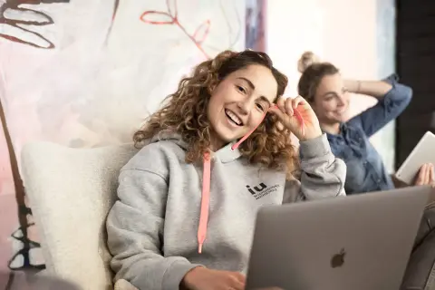 Zwei junge Frauen lernen entspannt. Eine trägt einen Hoodie mit dem Logo der IU Internationale Hochschule, die andere hält ein Tablet.