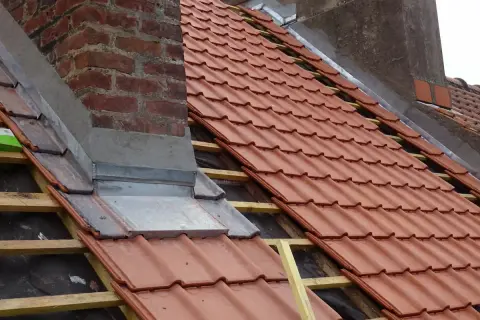 Dachdecker arbeiten am Schornstein, um ihn abzudichten. Neue rote Dachziegel werden verlegt.