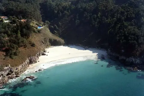 Vista aérea de la playa de San Pedro, Galicia, con aguas turquesas, arena blanca y vegetación exuberante en los acantilados.