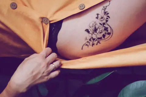 Ein zartes Tatoo mit einer Blume ziert die Haut, umrahmt von einem gelben Stoff und grünen Blättern.