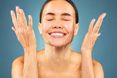Donna sorridente con acqua sul viso e mani. L'acqua micellare a cosa serve? A detergere e rinfrescare la pelle.