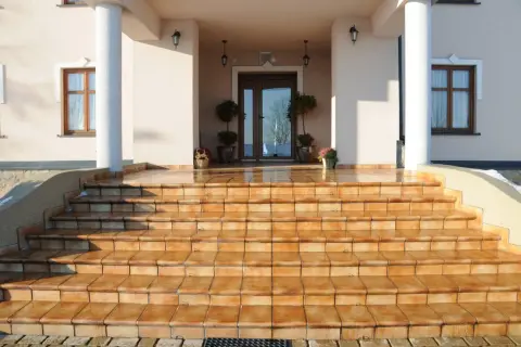 Amplia escalera abierta al exterior, con escalones de baldosas naranjas que conducen a una entrada principal con puerta doble y ventanas.