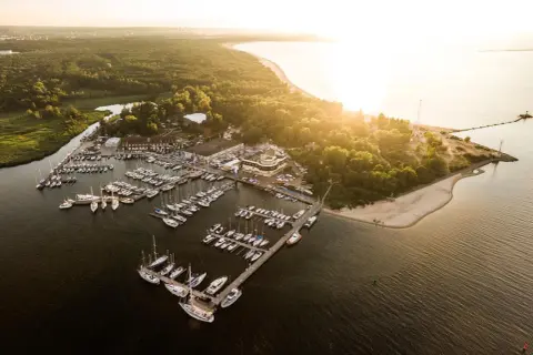 Zachód słońca nad zatoką, gdzie cumują liczne jachty. Widok na górki zachodnie marina, otoczoną zielenią i piaszczystą plażą.