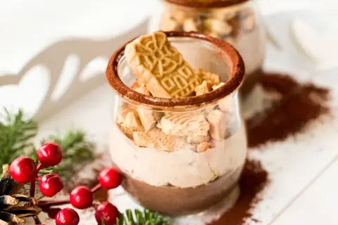 Zwei Gläser mit cremiger Spekulatius-Creme, garniert mit Spekulatius-Stücken und Kakaopulver. Weihnachtliche Deko mit Tannenzweigen und roten Beeren.