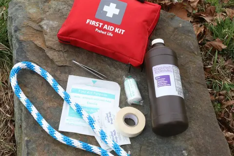 Contents of a DIY dog first aid kit laid out on a rock: red first aid bag, hydrogen peroxide, gauze, tape, alcohol pads, tweezers, and rope.