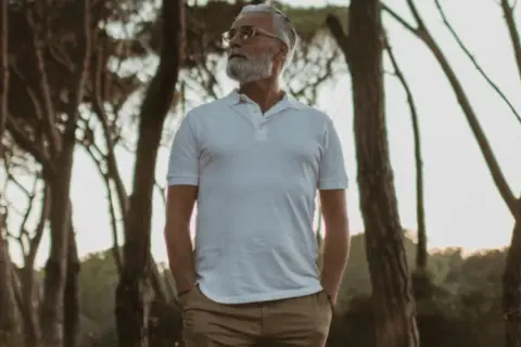 Älterer Mann mit Bart in weißem Polo und brauner Hose, steht im Wald. Die polo farben sind klassisch und zeitlos.