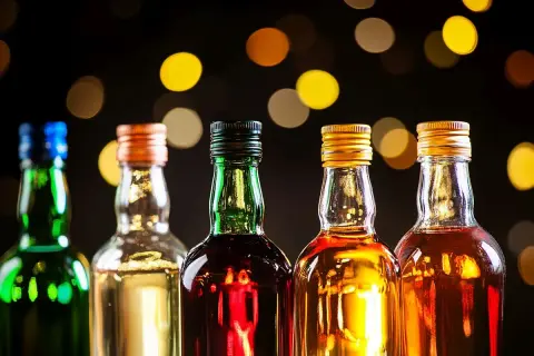 Rząd pięciu butelek z różnymi alkoholami, w tym zielona z niebieskim korkiem, przezroczysta z pomarańczowym, zielona z czarnym, przezroczysta ze złotym i przezroczysta z kolejnym złotym. Tło rozmyte, z ciepłymi, okrągłymi światłami. Idealne na wieczór ...