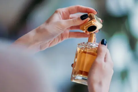 Dłonie z ciemnym lakierem do paznokci otwierają luksusowe perfumy. Złota nakrętka unosi się nad szklaną butelką z bursztynowym płynem, niczym zapowiedź zapachu Nasomatto Black Afgano.