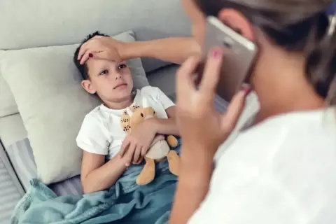 Chory chłopiec leży pod kocem, mama sprawdza mu temperaturę i dzwoni po pomoc. L4 na dziecko.