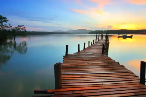 A wooden pier stretches into a calm lake at sunset, showcasing great depth of field photography. The sky transitions from blue to vibrant orange, reflecting on the water.