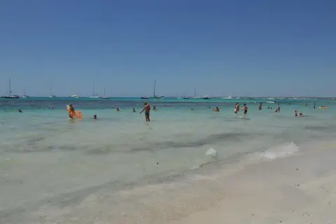 Słoneczny dzień na plaży Es Trenc. Ludzie kąpią się w turkusowej wodzie, a na horyzoncie widać żaglówki.