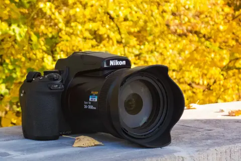 Czarny aparat Nikon P1000 z obiektywem na tle jesiennych, żółtych liści.