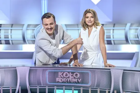 Uśmiechnięty mężczyzna i kobieta w białej sukience, którzy prowadzą Koło Fortuny, stoją przy pulpicie.