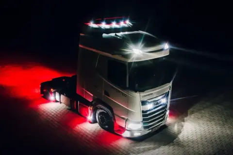 Camion DAF blanc illuminé par des feux de jour et des lumières rouges vives sur un sol pavé la nuit.