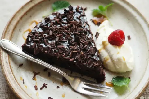 En bit glutenfri kladdkaka med chokladspån, vispad grädde, hallon och mynta på en tallrik.