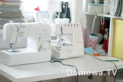 Domowy kącik do szycia z białą maszyną do szycia i owerlokiem. W tle stos materiałów i półki z akcesoriami.