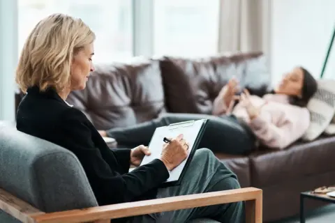 Una psicologa annota su un blocco note mentre una paziente sdraiata sul divano parla. La frequenza con cui si va dallo psicologo varia.