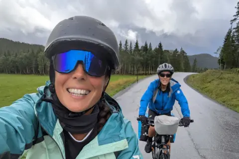 Dos ciclistas disfrutan de la ruta EuroVelo 6, con un paisaje verde y montañoso bajo un cielo nublado.