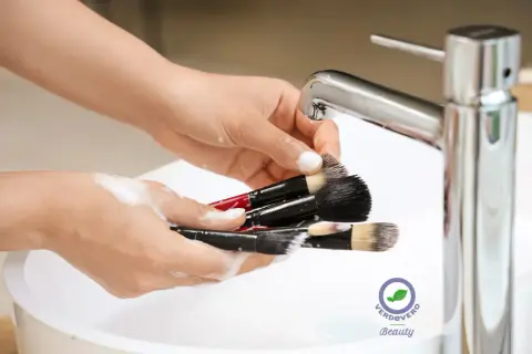 Mani che lavano pennelli da trucco insaponati sotto un rubinetto cromato. Un gesto di cura per un make-up impeccabile.