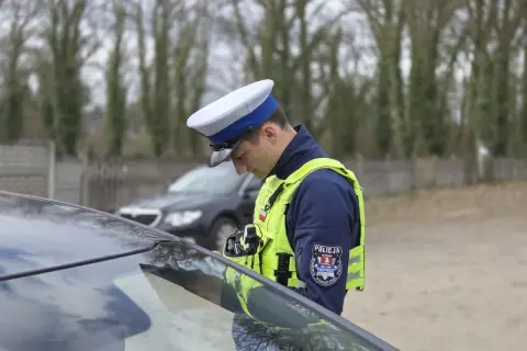 Policjant w kamizelce odblaskowej i czapce, pochylony nad maską samochodu, stosuje zasadę ograniczonego zaufania.