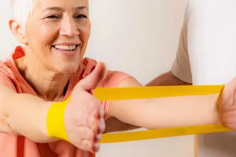 Uśmiechnięta starsza kobieta ćwiczy z fizjoterapeutą, używając żółtej gumy rehabilitacyjnej.