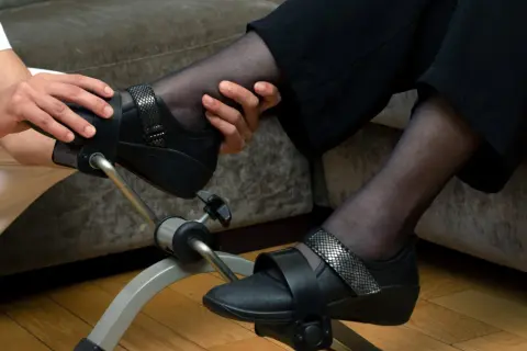 Osoba w białym stroju pomaga pacjentowi ćwiczyć na rowerku rehabilitacyjnym do wzmacniania mięśni rąk i nóg.