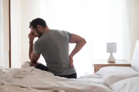 Homme assis sur un lit, main sur le front et l'autre sur le bas du dos, cherchant comment dormir avec une sciatique.
