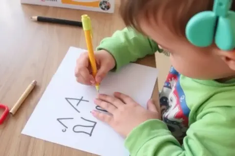 Dziecko z zielonymi słuchawkami ćwiczy pisanie, by pokonać dysgrafię. Na kartce widać litery.