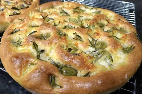 Une focaccia pizza dorée, garnie d'olives vertes et de romarin frais, sort du four.