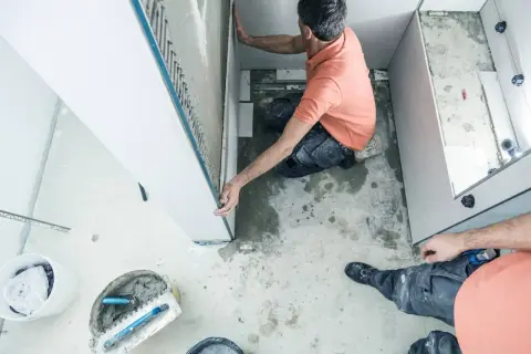 Un ouvrier réalise l'installation d'une douche, posant des panneaux muraux. Outils et matériaux de construction sont visibles au sol.