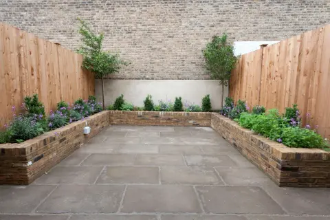 Un patio moderno con jardineras de ladrillo para decorar poyete ventana exterior. Plantas verdes y flores moradas crean un oasis urbano.