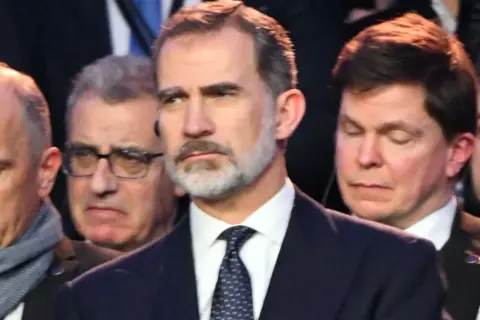 Król Hiszpanii Felipe VI w garniturze i krawacie, otoczony przez innych mężczyzn.
