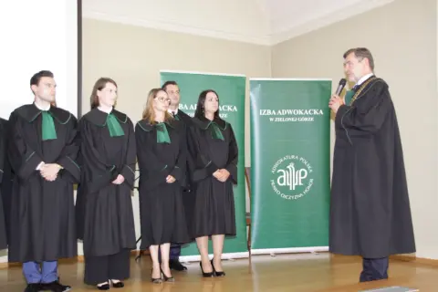 Grupa prawników w togach stoi przed zielonymi banerami Izby Adwokackiej.