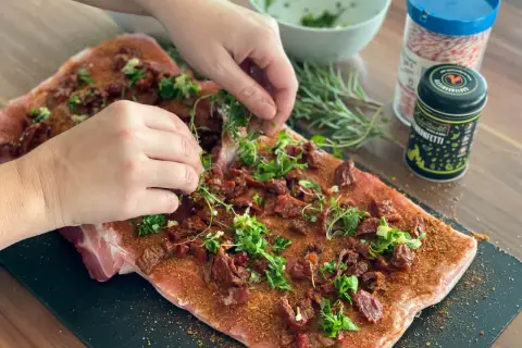 Hände belegen rohes Schweinefleisch mit Kräutern und getrockneten Tomaten für Porchetta.