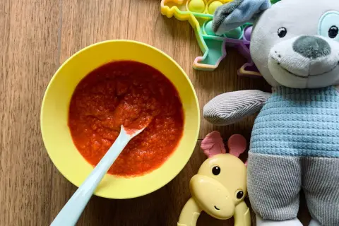 Pyszna zupa buraczkowa dla niemowlaka w żółtej miseczce, obok pluszowy piesek i gryzak.