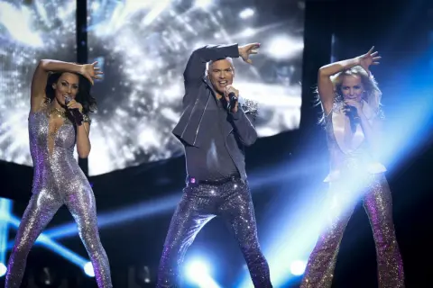 Yngves danskurser: Tre artister i glittriga scenkläder sjunger och dansar på en upplyst scen.