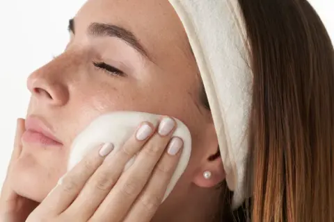 Mujer con diadema aplicándose espuma limpiadora en el rostro, parte de su rutina facial de noche.