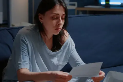 Zmartwiona kobieta czyta dokumenty, które mogą oznaczać eksmisję. Siedzi na kanapie, pogrążona w myślach.