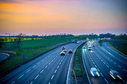 Autostrada z ruchem samochodowym o zachodzie słońca, z zielonymi polami w tle.