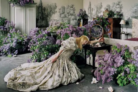 Una mujer rubia con un vestido estampado yace rodeada de lilas, una escena onírica capturada por Tim Walker.