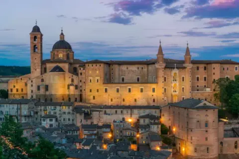Urbino Natale: il Palazzo Ducale illuminato al crepuscolo, con le sue torri e la cupola, domina la città storica.