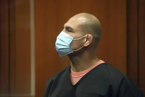 En skallig man med ljusblå mask bär en svart överdel över en rosa tröja. Harry Goularte ser ut att vara i en rättegångssal.